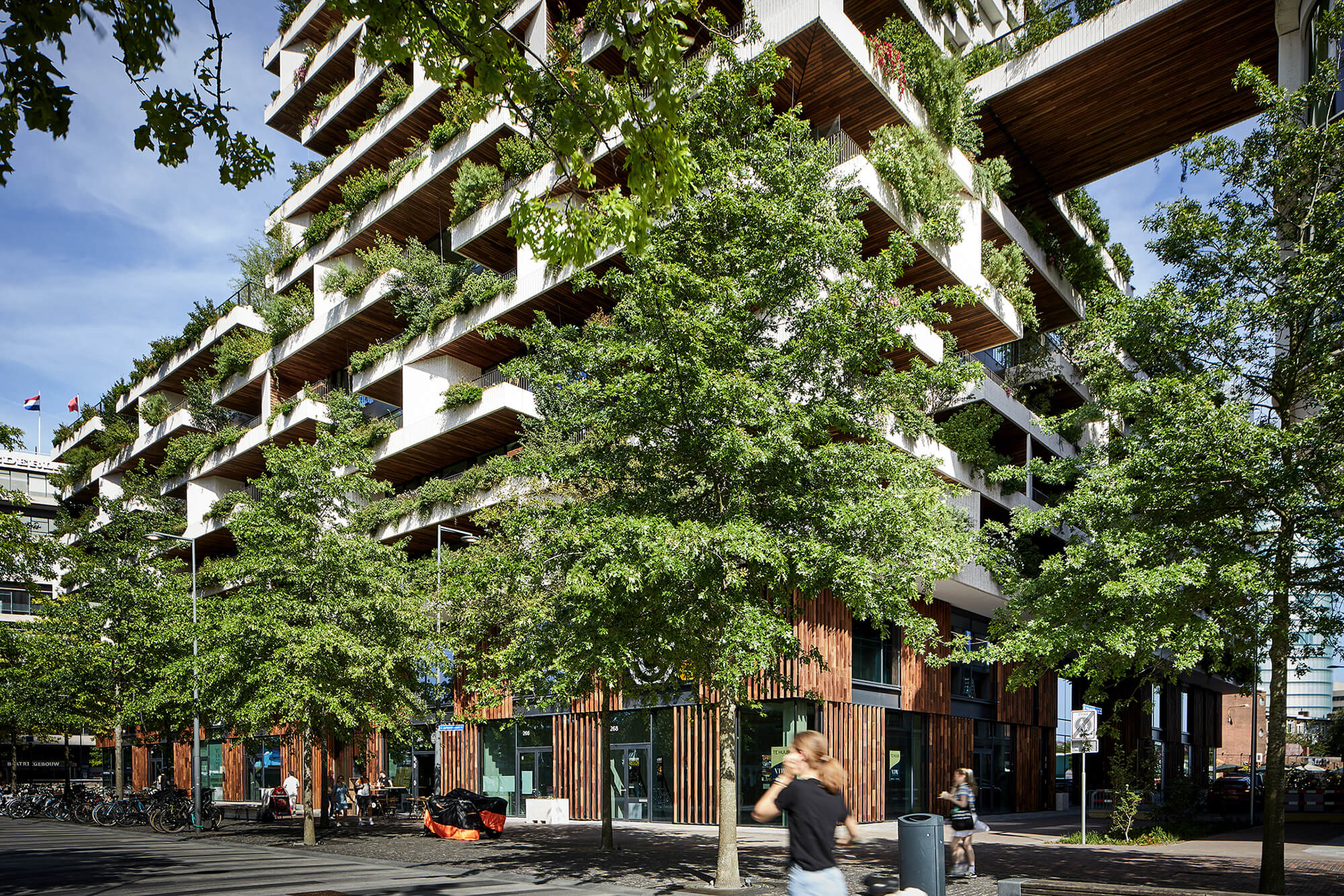 Fietsen en een voorbijganger bij de houten plint van Wonderwoods, met beplante balkons boven de winkelpuien.