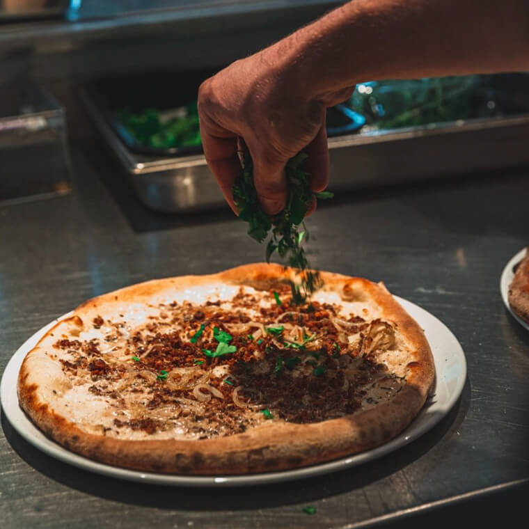 Kok bestrooit een pizza met verse kruiden in een restaurantkeuken.