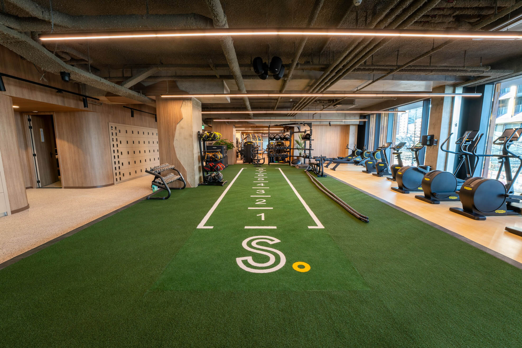 Fitnessruimte met een kunstgrasbaan met sprintmarkeringen en cardioapparatuur bij de ramen.